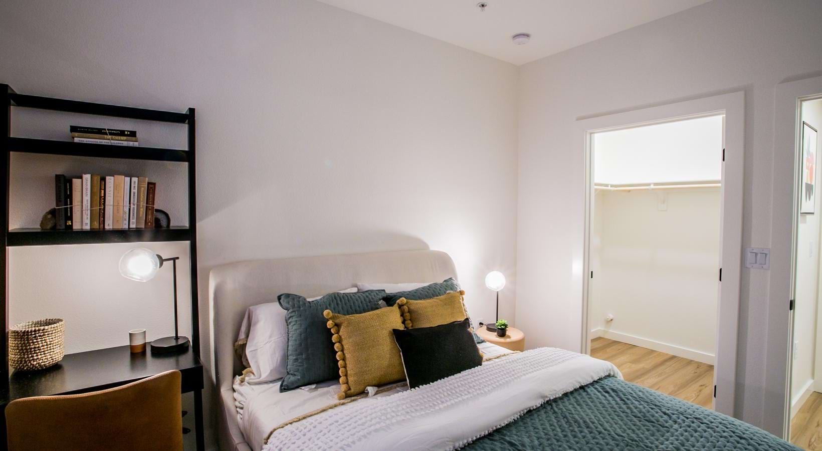A cozy apartment bedroom featuring a bed with decorative throw pillows, a dark desk, and an open closet doorway.