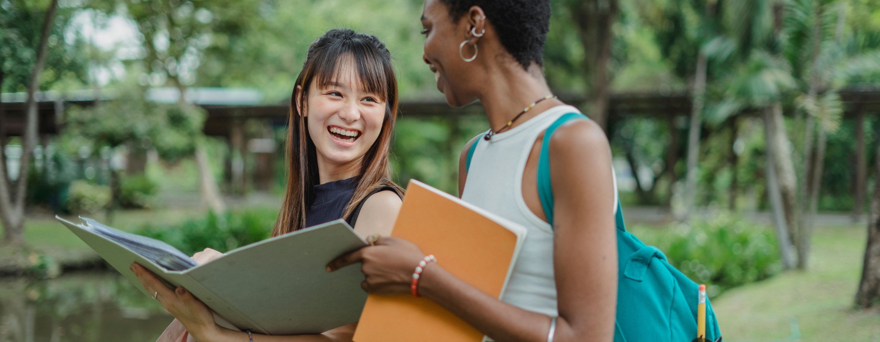 Two women standing outdoors in a park while holding a binder and an orange folder next to a body of water. 