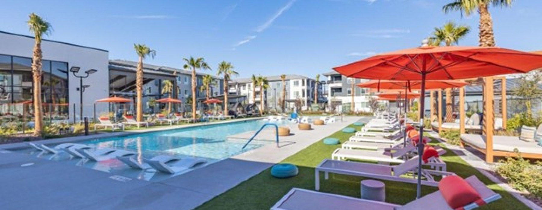 A resort-style apartment pool area featuring red umbrellas, lounge chairs, submerged seating, and palm trees.