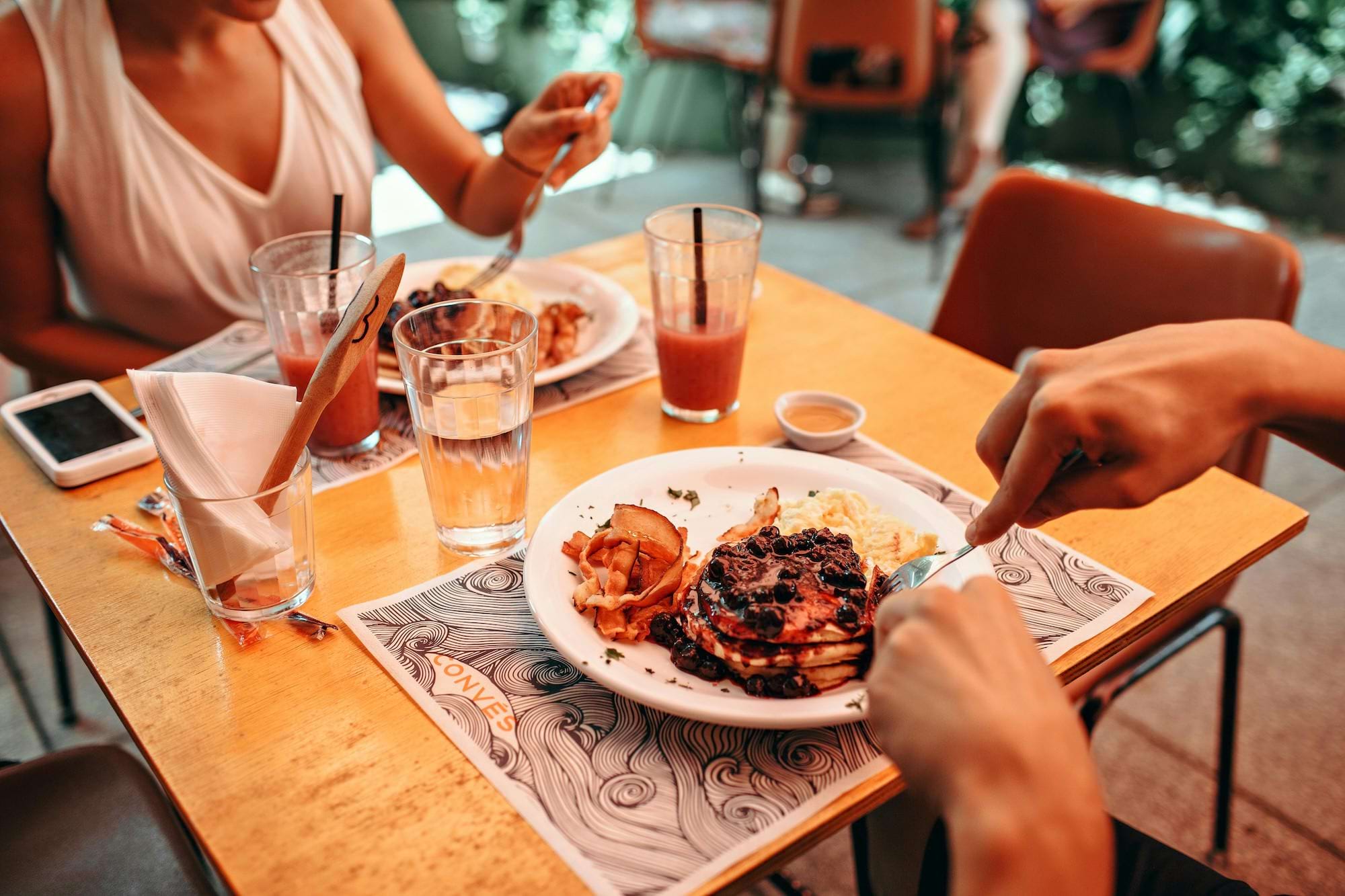 Two people eating pancakes and breakfast foods while sitting at a wooden dining table enjoying an outdoor meal.s while sitting at a wooden dining table enjoying an outdoor meal.